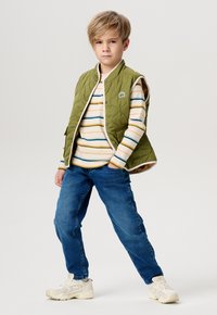 Grüne gesteppte Weste ohne Ärmel, getragen über einem cremefarbenen gestreiften Hemd mit blauen und orangefarbenen Linien, kombiniert mit blauen Jeans und hellen Sneakern.