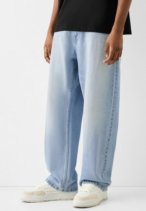 Persona che indossa jeans larghi azzurri, sneakers bianche e una maglietta nera, in piedi davanti a uno sfondo chiaro e semplice.