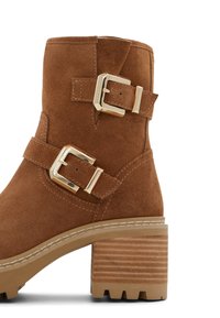 Botín de ante marrón con dos hebillas doradas, tacón de bloque de madera acanalada y suela de goma texturizada para mayor tracción.