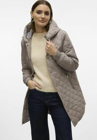 Gequilted beige jas met een capuchon, voorzien van drukknoopsluitingen en een rits aan de voorkant. Draag het over een lichtbeige trui en donkerblauwe jeans.