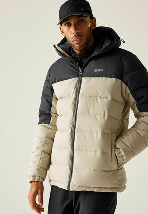 Mężczyzna w beżowo-czarnej puchowej kurtce z kapturem i czarnej czapce, stojący z jedną ręką w kieszeni na jednolitym tle.