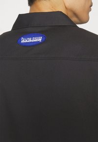 Maglietta nera con colletto corto, caratterizzata da una patch ricamata blu sulla parte posteriore che recita "FILLING PIECES DAGGER". Texture liscia e vestibilità standard.