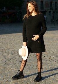 Robe noire en maille avec des poches, collants noirs à motifs, et bottines noires. Tient un chapeau blanc à large bord avec un bandeau beige.