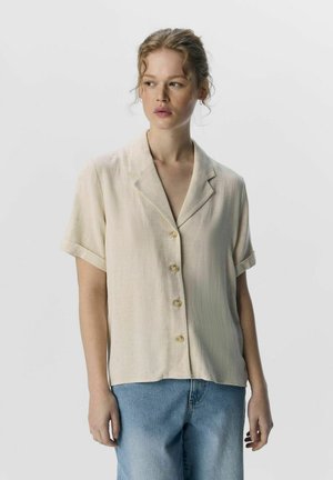 Chemise beige à manches courtes avec un col en V, présentant un tissage texturé et des boutons transparents, associée à un jean bleu clair.