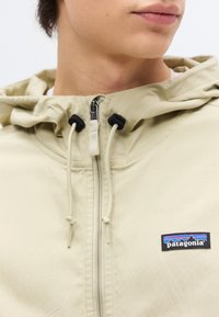 Νεαρό άτομο που φοράει μπεζ φούτερ με φερμουάρ και το λογότυπο της Patagonia στο στήθος, στραμμένο ελαφρώς στο πλάι μπροστά σε απλό φόντο.