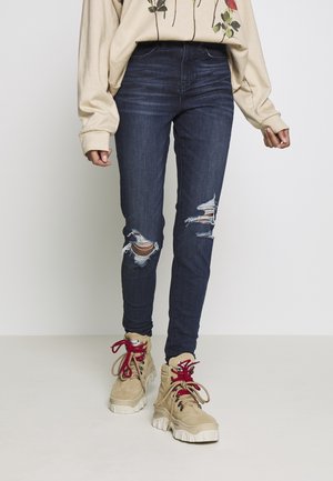 Vrouw draagt donkere gescheurde skinny jeans, beige oversized sweater met bloemenprint en beige laarzen met rode veters.