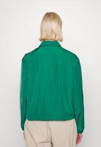 Chaqueta de nailon verde con cuello tipo solapa, puños elásticos y corte recto. Textura suave con rayas laterales en contraste.
