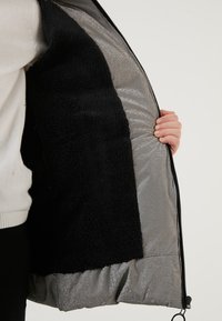 Sort fleeceforet jakke med en skinnende grå yderside, der har en lynlåslukning fortil og et rummeligt indre for varme. Synlig hånd holder stoffet.