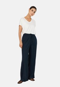 Bijela majica s v-neck izrezom u kombinaciji s tamnoplavim hlačama širokih nogavica koje imaju pojas s vezicom. Izgled je upotpunjen smeškim sandalama s otvorenim prstima.