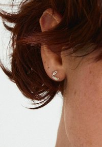 Pendiente de plata con un diseño curvado, que presenta piedras preciosas de colores: rosa, verde y transparente. Lleva puesto en el lóbulo de una oreja con cabello corto de color castaño rojizo.