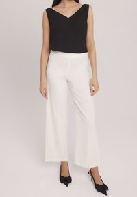 Top negro sin mangas con escote en V; pantalones blancos de pierna ancha; tacones negros de punta con detalle de lazo. Tejidos suaves, diseño moderno.