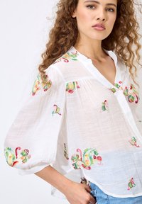 White, lightweight blouse with colorful floral embroidery in red, green, and yellow. Features a collared neckline and billowy sleeves.
