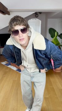 Ναυτική βομβιστική τζάκετ με μπεζ γούνα στον γιακά, γκρι φούτερ, ανοιχτό γκρι τζιν και σκούρα γυαλιά ηλίου, που στέκεται σε ξύλινο πάτωμα.