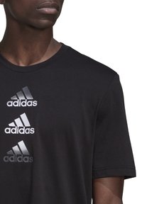 Muž v černém tričku s třemi vertikálními logy Adidas v bílé, stříbrné a černé barvě na levé přední straně.