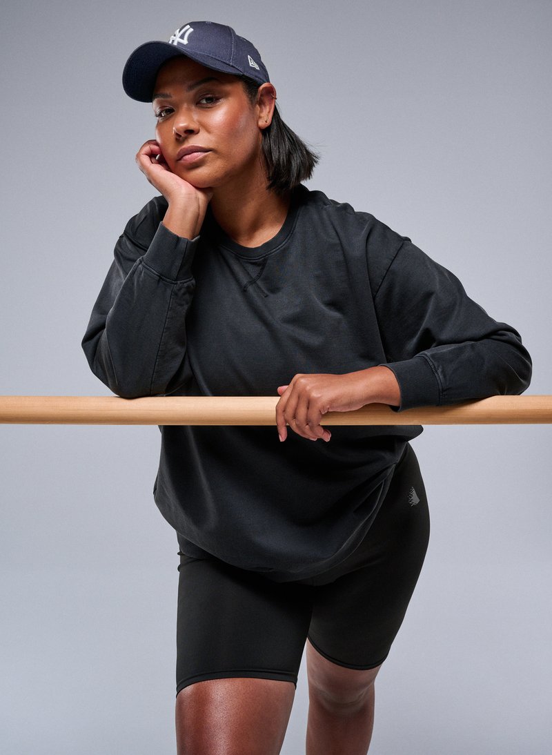 Felpa nera oversize e pantaloni corti neri da ciclista. La modella si appoggia a una barra di legno, indossando un berretto blu navy con un logo bianco. Sfondo neutro.
