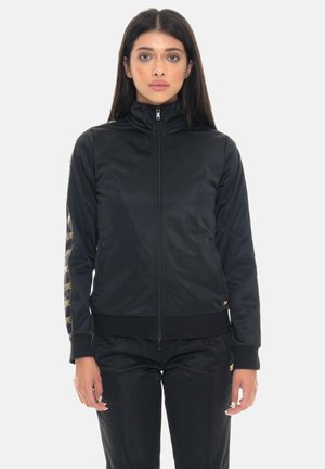 Junge Frau mit langen Haaren, die eine schwarze Jacke mit Reißverschluss und passende schwarze Hose trägt, steht vor einem schlichten weißen Hintergrund.