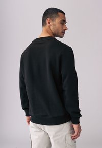 Črna sweatshirt s okroglo ovratnico, rebrastimi manšetami in robom; izdelana iz mehkega materiala, ki prikazuje preprost, čist dizajn.