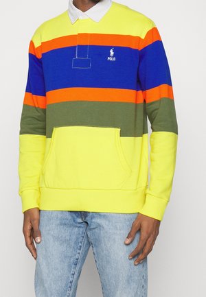 Homme portant un polo à manches longues avec des rayures horizontales jaunes, oranges, bleues et vertes et un jean bleu clair.