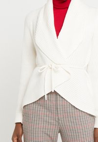 Jersey blanco con pliegues y un amplio cuello, atado con un cinturón, superpuesto sobre un suéter de cuello alto rojo, combinado con pantalones de cuadros en tonos neutros.