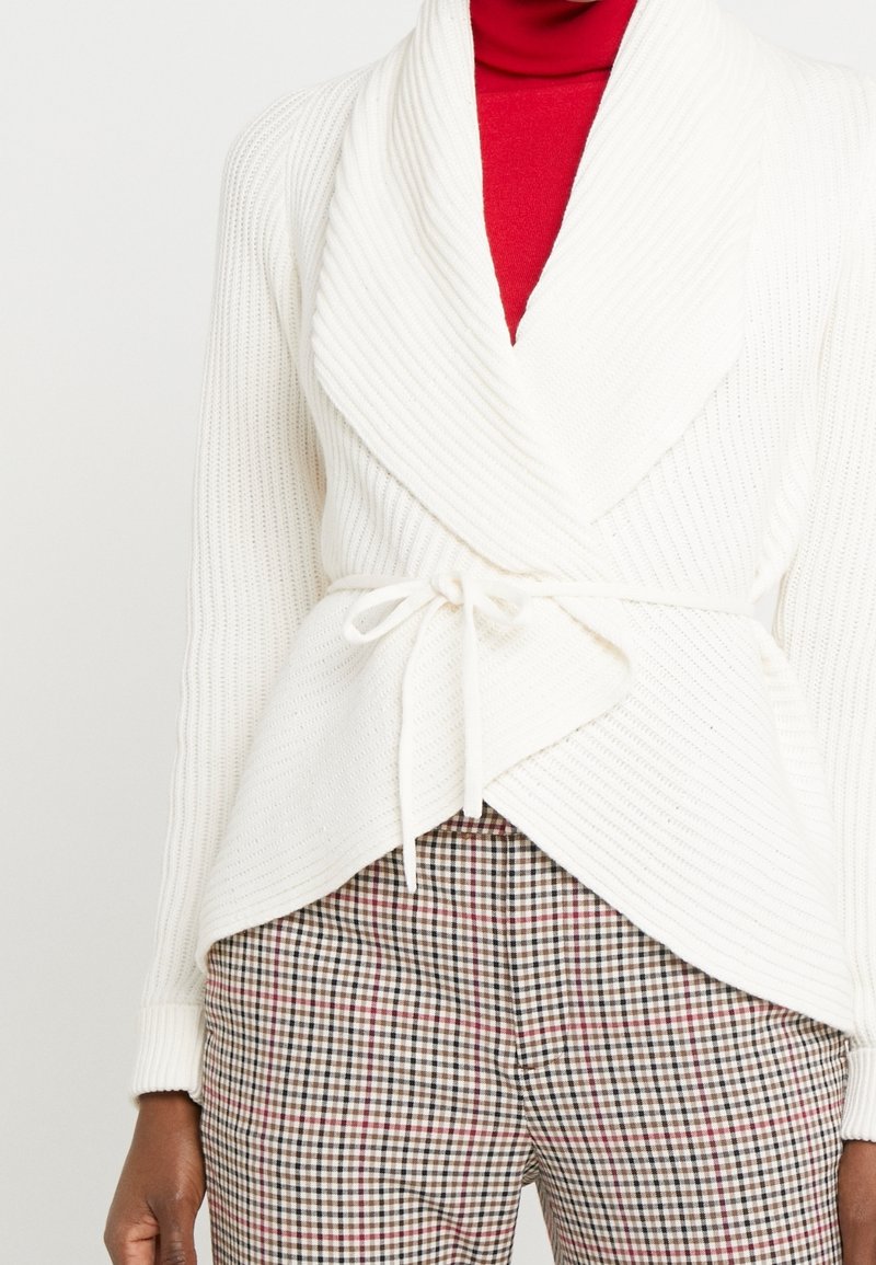 Jersey blanco con pliegues y un amplio cuello, atado con un cinturón, superpuesto sobre un suéter de cuello alto rojo, combinado con pantalones de cuadros en tonos neutros.