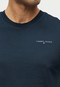 Άνδρας που φοράει μπλε ναυτικό T-shirt με στρογγυλή λαιμόκοψη και μικρό λευκό κείμενο "Tommy Jeans" και λογότυπο στο αριστερό μέρος του στήθους.