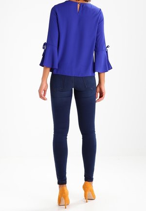 Femme portant un chemisier bleu cobalt à manches évasées et poignets noués, un jean skinny foncé et des chaussures à talons hauts jaune moutarde, vue de dos.