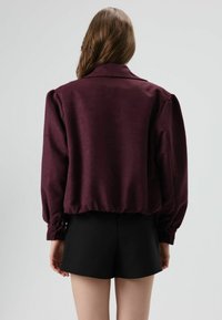 Bordeauxkleurige satijnen blouse met een kraag, pofmouwen en een elastische zoom. Gestyled met zwarte korte broek. Soepele textuur en aansluitend ontwerp.