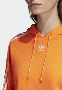 Oranžová mikina s kapucí disponující klokankou, bílým designem se třemi pruhy na rukávech a vyšitým logem na hrudi.