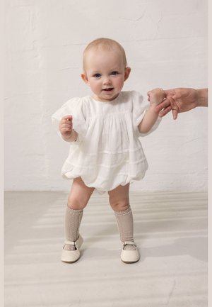 Fehér ruhába öltözött kislány bézs térdzokniban, aki egy felnőtt kezét fogja, miközben áll egy egyszerű világos háttér előtt.