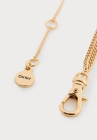 Lănțișor de aur cu închidere tip homar și etichetă rotundă gravată cu "DKNY" pe un fundal alb.