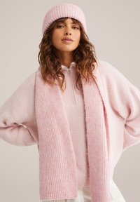 Rozā adīta džemperis ar augstu kakla rāvējslēdzēju, apvienots ar atbilstošu ribotu šalli un beanie cepuri, visiem piemīt maiga, teksturēta apdare.