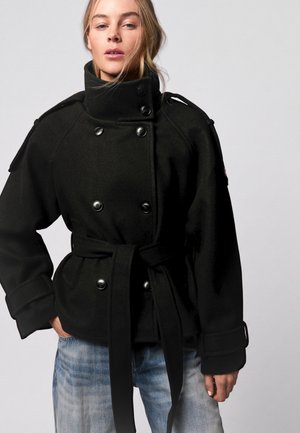 Lyshåret kvinde iført en sort dobbeltradet frakke med høj krave og bælte, kombineret med lyseblå jeans, stående mod en ensfarvet baggrund.