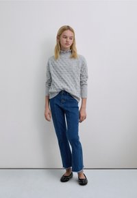 Grijze gebreide trui met een hoge hals, gecombineerd met blauwe high-waisted jeans met een knoopsluiting aan de voorkant en rafelige zomen, en zwarte platte schoenen met studs.