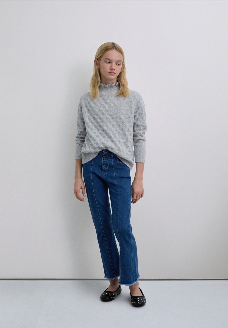 Grijze gebreide trui met een hoge hals, gecombineerd met blauwe high-waisted jeans met een knoopsluiting aan de voorkant en rafelige zomen, en zwarte platte schoenen met studs.