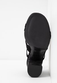Semelle de sandale à talon noire avec grip texturé, tenue debout contre un mur blanc d'angle.