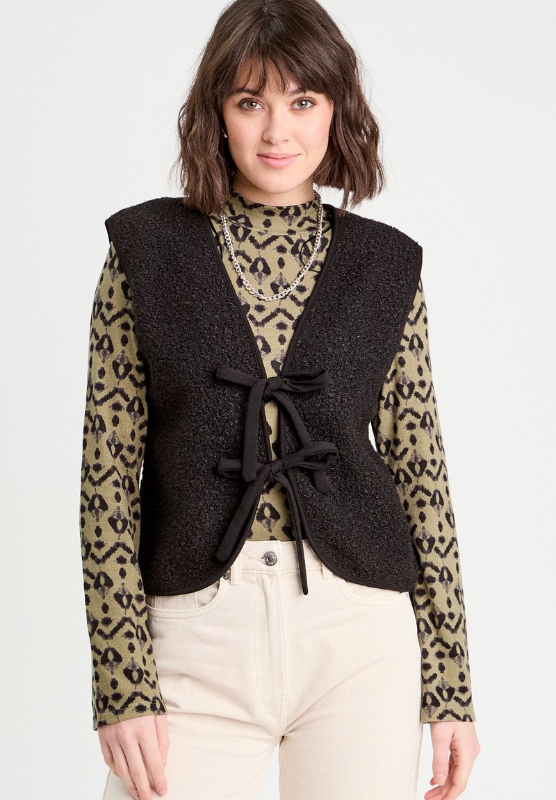 Gilet nero texture con chiusura a cravatta, indossato sopra un dolcevita verde con motivo. Pantaloni chiari completano l'outfit.