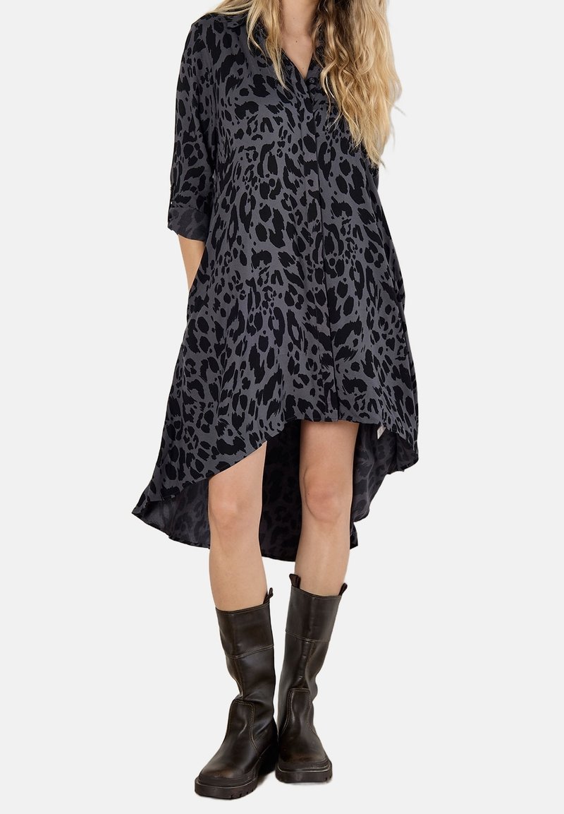 Femme portant une robe noire et grise à imprimé léopard asymétrique, manches trois-quarts et bottes noires mi-mollet, mains dans les poches.