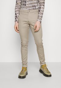Beige slim-fit housut, jotka on valmistettu puuvillasekoituksesta ja joissa on etutaskut sekä nappikiinnitys, yhdistettynä oliivinvihreisiin saappaisiin, joissa on keltaiset nauhat.