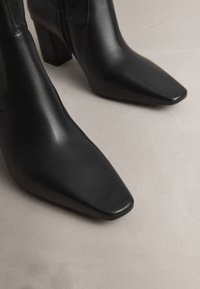 Svarta läderankelstövlar med spetsig tå, fyrkantig klack och slät textur, utrustade med en dragkedja längs sidan.
