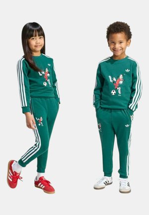 Due bambini indossano tute Adidas verdi coordinate con strisce bianche e un disegno di un giocatore di calcio dei cartoni animati, uno con scarpe rosse e l'altro con scarpe bianche.