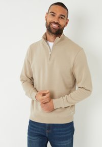 Sweatshirt beige à zip quart avec des poignets et une taille côtelés. Texture douce, coupe décontractée, avec une petite étiquette de marque en bas.