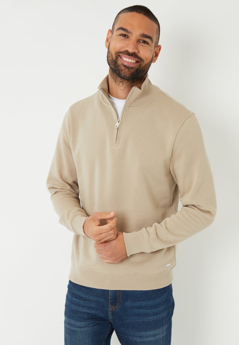 Sweatshirt beige à zip quart avec des poignets et une taille côtelés. Texture douce, coupe décontractée, avec une petite étiquette de marque en bas.