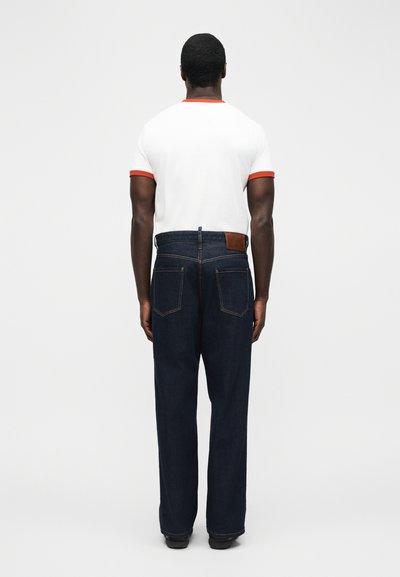 Άνδρας που στέκεται με την πλάτη γυρισμένη, φορώντας λευκό T-shirt με πορτοκαλί περίγραμμα και σκουρό μπλε τζιν με ίσια γραμμή, πίσω τσέπες και δερμάτινη ετικέτα.