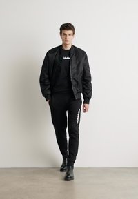 Crna bomber jakna preko crne majice i crnih trenirki, s bijelim logotipom. Model nosi crne čizme, stojeći na betonskom podu.
