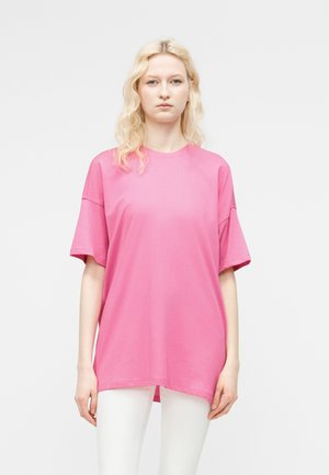 Blonde vrouw met golvend haar staat recht voor zich uit kijkend, draagt een losse felroze shirt met korte mouwen en witte broek tegen een lichte achtergrond.