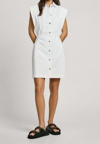 Biała sukienka bez rękawów z przodem na guziki i dopasowaną talią. Uzupełniona czarnymi sandałami z ćwiekami i grubą podeszwą.