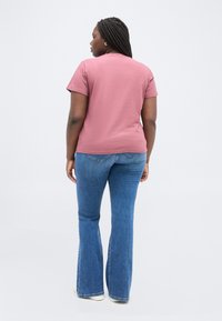 Γυναίκα με πλεξούδες που φοράει ροζ μπλουζάκι με κοντά μανίκια, μπλε τζιν παντελόνι με φαρδιά ποδόγυρα και λευκά παπούτσια, στέκεται γυρισμένη προς τα πίσω με φόντο λευκό.