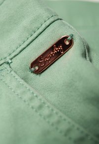 Lichtgroene stof met een gladde textuur, voorzien van een metalen label met de gravure "Superdry". Zichtbare stikseldetails langs de rand.