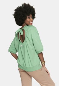 Miętowa bluza z krótkimi rękawami, projekt z otwartych pleców związany w kokardę, teksturowana tkanina i dopasowany pas. Widać beżowe spodnie.