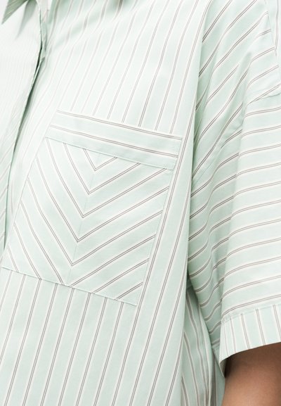 Ανοιχτό μπλε πουκάμισο με λεπτές κάθετες και ζικ ζακ ρίγες στην τσέπη, κοντό μανίκι ορατό στη δεξιά πλευρά, το ύφασμα φαίνεται μαλακό και λείο.